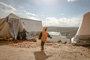 Anonymous boy carrying heavy plastic canister while poor girl watching near tents under cloudy sunny sky